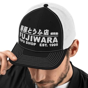 May include: Black and white trucker hat with the text "FUJIWARA TOFU SHOP EST. 1995" in white. The hat has a black front panel and bill, with a white mesh back. The hat is being held by a person.