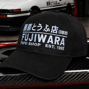 May include: A black baseball cap featuring white embroidered text that reads Fujiwara Tofu Shop Est. 1995 along with Japanese characters. The cap has a structured crown and a curved brim, displaying a clean design with high-contrast white lettering.