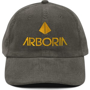 May include: A dark gray corduroy baseball cap with the word "ARBORIA" embroidered in gold thread. Above the word is a gold geometric design. The cap has a curved brim and an adjustable strap.