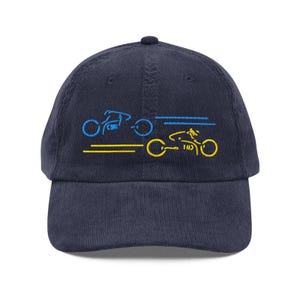 May include: Navy blue corduroy baseball cap with embroidered yellow and blue bicycle designs. The cap features a curved brim and a fabric-covered adjustable strap at the back. The embroidered designs depict two bicycles in motion.