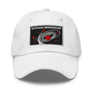 Puede incluir: Una gorra de b&eacute;isbol blanca con un parche rectangular bordado en la parte delantera. El parche tiene un fondo negro con el texto blanco GOTHAM OBSERVATORY sobre un gr&aacute;fico de una galaxia espiral blanca, un planeta rojo y estrellas rojas.