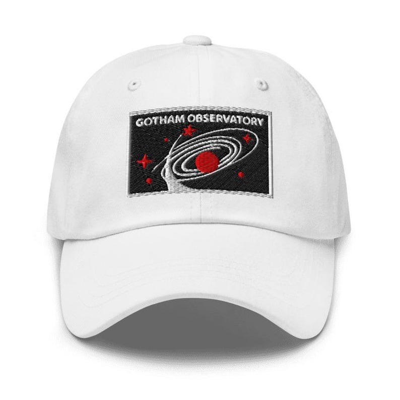 Puede incluir: Una gorra de b&eacute;isbol blanca con un parche rectangular bordado en la parte delantera. El parche tiene un fondo negro con el texto blanco GOTHAM OBSERVATORY sobre un gr&aacute;fico de una galaxia espiral blanca, un planeta rojo y estrellas rojas.