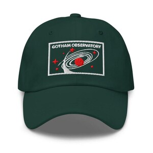 Puede incluir: Una gorra de b&eacute;isbol de color verde oscuro con un parche rectangular bordado en la parte delantera. El parche muestra el texto GOTHAM OBSERVATORY en blanco, con un planeta rojo, anillos orbitales blancos y peque&ntilde;as estrellas rojas.