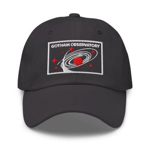 Puede incluir: Una gorra de b&eacute;isbol negra con un parche rectangular bordado en la parte delantera. El parche muestra el texto GOTHAM OBSERVATORY en letras blancas sobre un gr&aacute;fico de un planeta rojo rodeado de anillos orbitales blancos y estrellas.