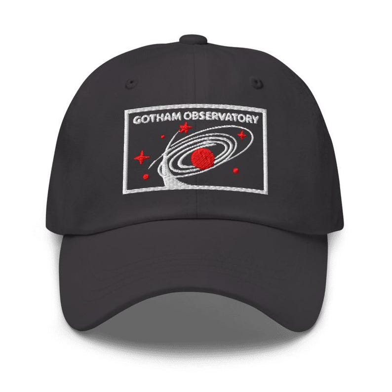 Puede incluir: Una gorra de b&eacute;isbol negra con un parche rectangular bordado en la parte delantera. El parche muestra el texto GOTHAM OBSERVATORY en letras blancas sobre un gr&aacute;fico de un planeta rojo rodeado de anillos orbitales blancos y estrellas.