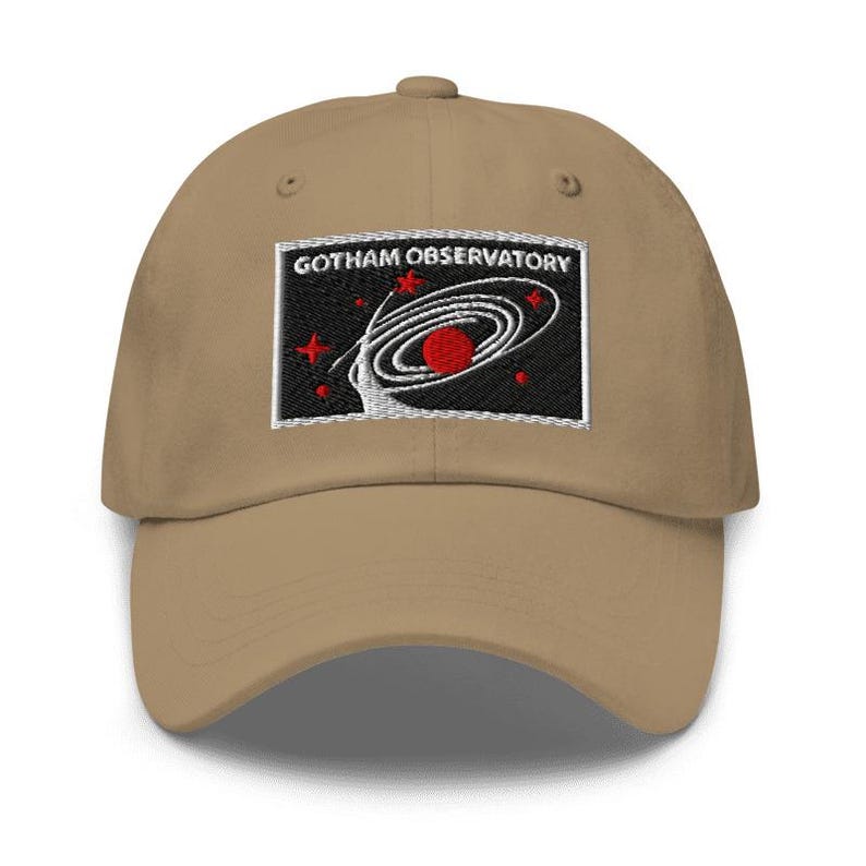 Puede incluir: Una gorra de b&eacute;isbol de color tostado con un parche rectangular bordado en la parte delantera. El parche tiene un fondo negro con el texto blanco GOTHAM OBSERVATORY sobre un gr&aacute;fico de galaxia espiral blanca, un planeta rojo y estrellas.