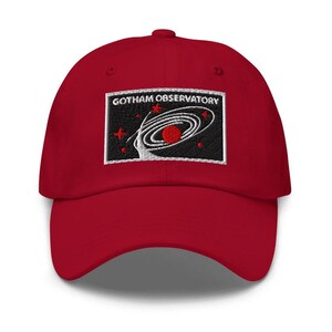 Puede incluir: Una gorra de b&eacute;isbol roja con un parche rectangular bordado en la parte delantera. El parche tiene un fondo negro con el texto blanco GOTHAM OBSERVATORY sobre un gr&aacute;fico de galaxia espiral blanca, un planeta rojo y varias estrellas rojas.