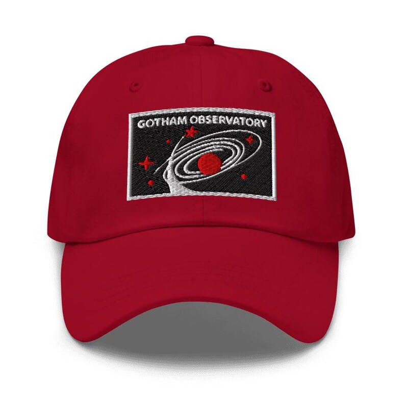 Puede incluir: Una gorra de b&eacute;isbol roja con un parche rectangular bordado en la parte delantera. El parche tiene un fondo negro con el texto blanco GOTHAM OBSERVATORY sobre un gr&aacute;fico de galaxia espiral blanca, un planeta rojo y varias estrellas rojas.