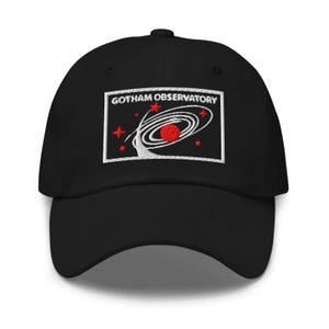Puede incluir: Una gorra de b&eacute;isbol negra con un parche rectangular bordado en la parte delantera. El parche muestra el texto GOTHAM OBSERVATORY en letras blancas sobre un gr&aacute;fico de un planeta rojo rodeado por anillos orbitales blancos y estrellas.
