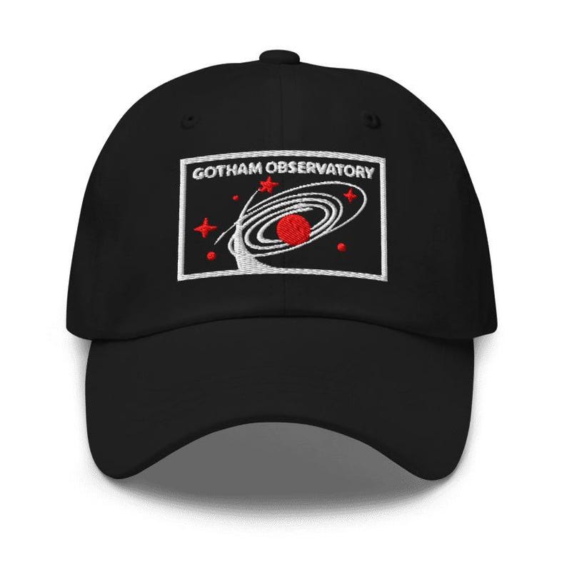 Puede incluir: Una gorra de b&eacute;isbol negra con un parche rectangular bordado en la parte delantera. El parche muestra el texto GOTHAM OBSERVATORY en letras blancas sobre un gr&aacute;fico de un planeta rojo rodeado por anillos orbitales blancos y estrellas.