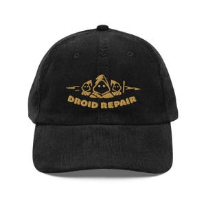 May include: Black corduroy baseball cap with a curved brim. The cap features a gold embroidered design of three hooded figures and the words "DROID REPAIR" in gold lettering. The cap has a structured fit and a top button.