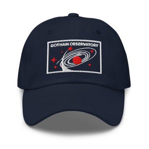 Puede incluir: Una gorra de b&eacute;isbol azul marino con un parche rectangular bordado en la parte delantera. El parche muestra el texto GOTHAM OBSERVATORY en letras blancas sobre un gr&aacute;fico de un planeta rojo rodeado de anillos orbitales blancos y estrellas.