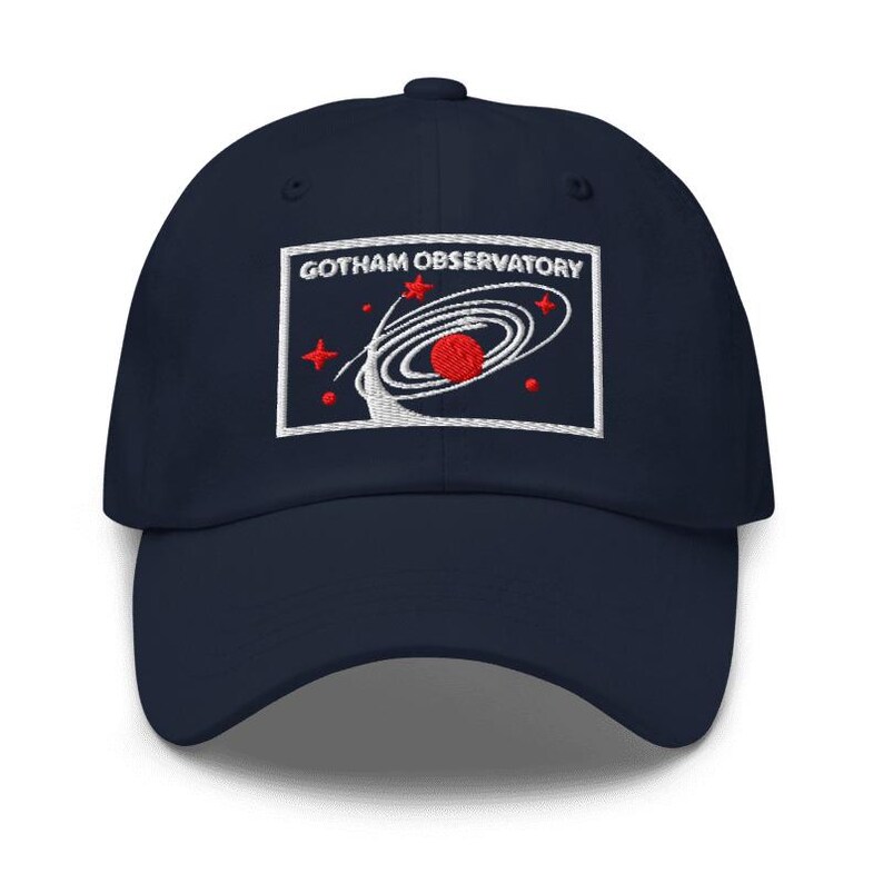Puede incluir: Una gorra de b&eacute;isbol azul marino con un parche rectangular bordado en la parte delantera. El parche muestra el texto GOTHAM OBSERVATORY en letras blancas sobre un gr&aacute;fico de un planeta rojo rodeado de anillos orbitales blancos y estrellas.