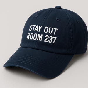 May include: A navy blue baseball cap featuring the embroidered text "STAY OUT ROOM 237" in white. The cap has a curved peak and a button on top. The hat is made of a sturdy fabric.