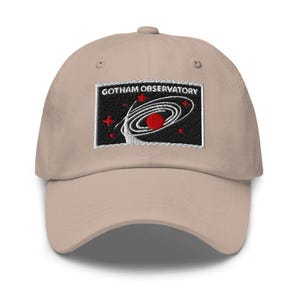 Puede incluir: Una gorra de b&eacute;isbol beige con un parche rectangular bordado en la parte delantera. El parche tiene un fondo negro con el texto blanco GOTHAM OBSERVATORY sobre un gr&aacute;fico de galaxia espiral blanca, un planeta rojo y varias estrellas rojas.