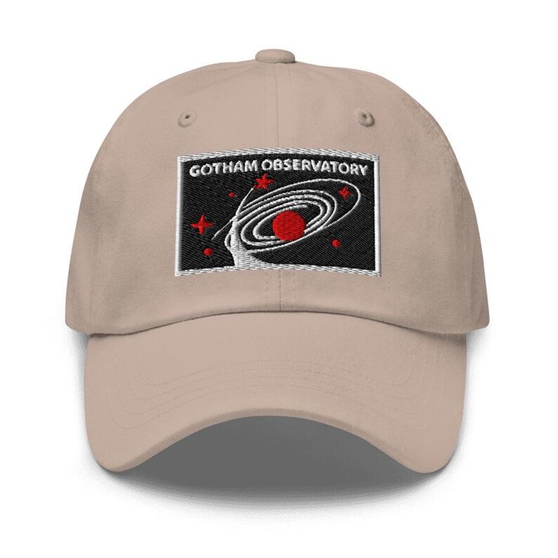 Puede incluir: Una gorra de b&eacute;isbol beige con un parche rectangular bordado en la parte delantera. El parche tiene un fondo negro con el texto blanco GOTHAM OBSERVATORY sobre un gr&aacute;fico de galaxia espiral blanca, un planeta rojo y varias estrellas rojas.