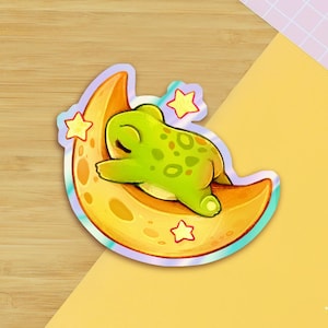 Peut inclure: Un autocollant représentant une grenouille de dessin animé dormant sur un croissant de lune. La grenouille est verte avec des taches vert foncé. La lune est jaune et orange avec trois étoiles jaunes. L'autocollant a une bordure holographique.