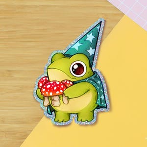 May include: A whimsical sticker featuring a green frog wearing a wizard hat and cape, holding red and white spotted mushrooms. The sticker has a holographic border and is set against a wooden background.
