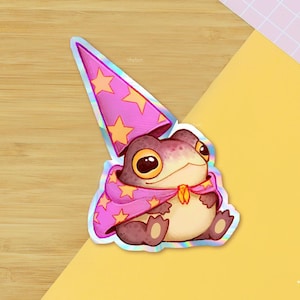 Può includere: Un adesivo stravagante con un ranocchio dei cartoni animati vestito da mago. Il ranocchio indossa un cappello e un mantello rosa adornati da stelle dorate. L'adesivo ha una finitura olografica ed è ambientato su uno sfondo di legno e giallo.