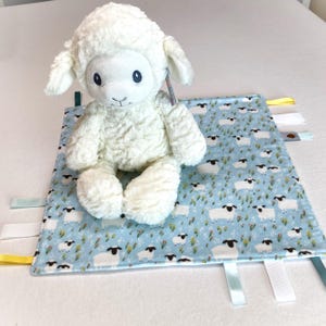 以下が含まれることがあります： 白い羊のぬいぐるみは、羊の模様が入った青いブランケットの上に置かれています。ブランケットの端にはカラフルなリボンが付いています。羊は柔らかく、質感のある外観で、濃い青色の目と小さな笑顔をしています。
