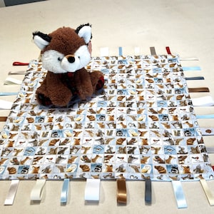 May include: A white baby blanket with a variety of animal faces printed on it. The blanket has multiple blue, brown, and white satin ribbons attached to the edges. A brown plush fox toy is sitting on top of the blanket.