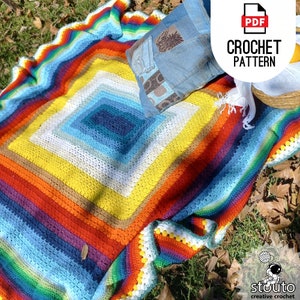 Puede incluir: Una manta de crochet colorida con un patrón de arcoíris. La manta está colocada sobre una superficie cubierta de hierba con una almohada y una cesta en el fondo. El texto "CROCHET PATTERN" es visible en la esquina superior derecha de la imagen.