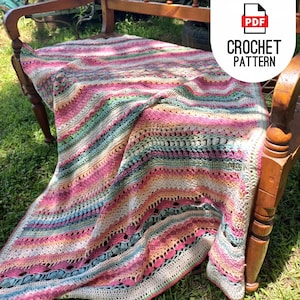 Op de afbeelding: Een gehaakt deken met een gestreept patroon in tinten roze, perzik, groen en blauw. Het deken is over een houten bank gelegd.