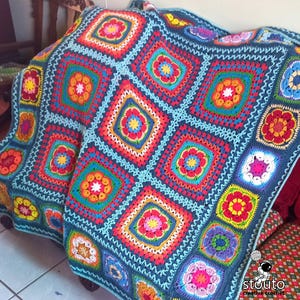 Könnte beinhalten: Eine bunte Häkeldecke mit blauem Hintergrund und einem sich wiederholenden Muster aus mehrfarbigen Blumen. Die Decke ist aus verschiedenen Garnfarben gefertigt, darunter Rot, Orange, Gelb, Grün, Blau und Lila.