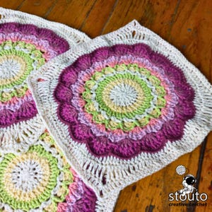 May include: Two crocheted squares with a mandala design in shades of pink, green, and cream. The squares are joined together and have a white border.