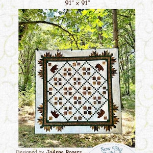 Op de afbeelding: Een handgemaakte quilt, getiteld "Kodiak Makin' Tracks", meet 231 cm x 231 cm. De quilt heeft een centraal patroon van bruine en groene vierkanten met berensilhouetten. De rand is donkergroen met bladvormen. De tekst "Sew Big Quilting" is ook zichtbaar.