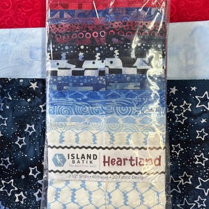 May include: A package of fabric strips in shades of blue, red, and white, with various patterns. The package is labeled "Heartland" and contains 2 1/2 inch strips, 40 pieces, and 20 fabric designs. The background features red, light blue, and dark blue fabrics.