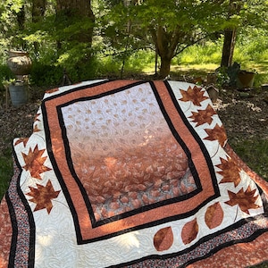 May include: A handmade quilt with a fall theme. The quilt features a central design of falling leaves in shades of orange and brown, framed by a black and rust-colored border. The corners and edges are adorned with leaf appliqués and floral patterns.