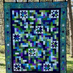 May include: A rectangular quilt featuring a patchwork design of blue, green, and purple batik fabric squares. The quilt includes eight white star-shaped blocks arranged in a grid, surrounded by a dark blue inner border and a mottled green outer border.