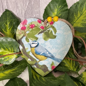 May include: A bluebird with yellow accents sits on a branch in a heart-shaped wooden ornament. The ornament is decorated with a floral pattern and a blue and white lace design.