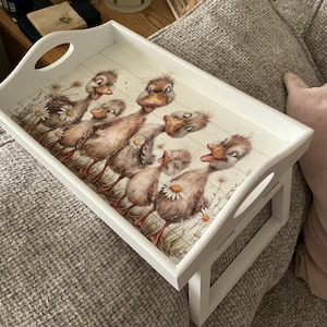 May include: A white wooden serving tray featuring a charming illustration of ducklings and daisies. The tray has a rectangular shape with rounded corners and handles. The ducklings are depicted in shades of brown and tan.