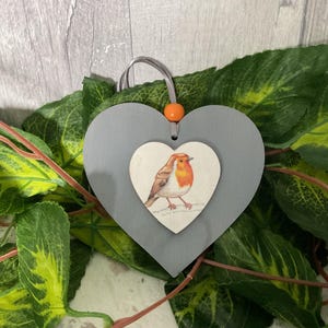 May include: A grey wooden heart-shaped ornament with a white heart cutout featuring a painted robin. The robin is orange, white, and brown. The ornament has a small orange bead at the top.
