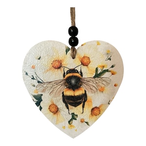 May include: Heart-shaped wooden ornament with a bumblebee and floral design. The bee is black and yellow, surrounded by yellow and white flowers. It hangs from a twine cord with two black beads. The background has a textured, off-white finish.