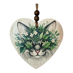 May include: Wooden heart-shaped ornament depicting a cat's face with a floral crown. The cat has green eyes, a pink nose, and fluffy white and grey fur. It hangs from a twine string with two wooden beads. The ornament is a decorative item.