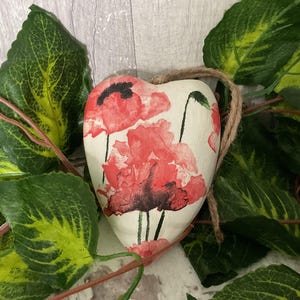 May include: White heart-shaped ornament with a floral design featuring red poppies. The ornament is surrounded by green leaves.