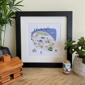 Puede incluir: Impresi&oacute;n art&iacute;stica enmarcada de Edimburgo, Escocia, con una colorida ilustraci&oacute;n de mapa con puntos de referencia. La impresi&oacute;n est&aacute; en un marco negro con un passe-partout blanco, exhibida con plantas en macetas y una figura de caba&ntilde;a de madera.