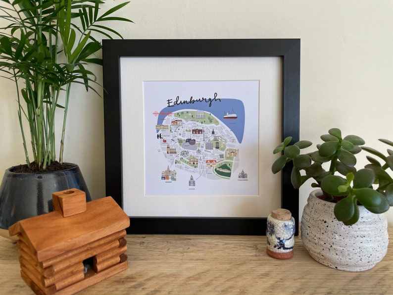 Puede incluir: Impresi&oacute;n art&iacute;stica enmarcada de Edimburgo, Escocia, con una colorida ilustraci&oacute;n de mapa con puntos de referencia. La impresi&oacute;n est&aacute; en un marco negro con un passe-partout blanco, exhibida con plantas en macetas y una figura de caba&ntilde;a de madera.