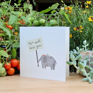 May include: A white card with a gray elephant holding a sign that says "we will miss you!" The card is sitting on a wooden surface with green plants in the background.