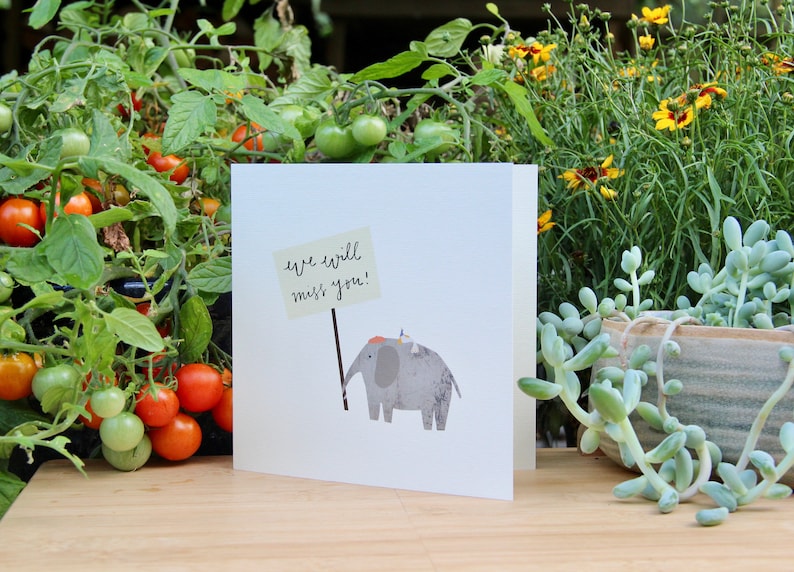 May include: A white card with a gray elephant holding a sign that says "we will miss you!" The card is sitting on a wooden surface with green plants in the background.