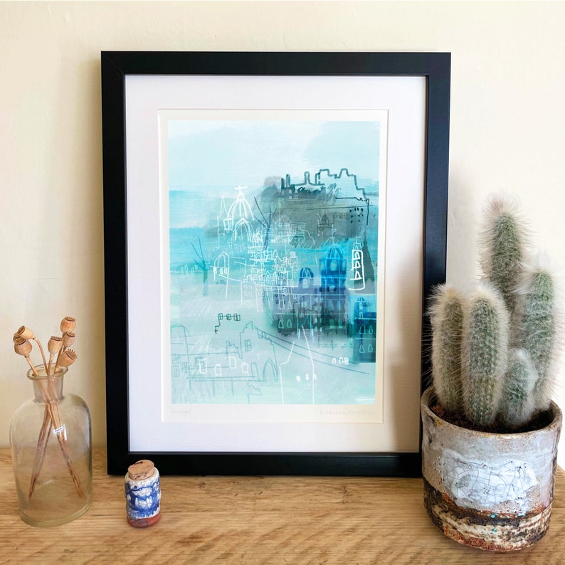 Puede incluir: Una impresi&oacute;n art&iacute;stica de paisaje urbano abstracto en un marco negro. La obra de arte presenta un estilo acuarela con tonos azules y grises, que representa edificios y detalles arquitect&oacute;nicos. Tambi&eacute;n se ve un cactus en una maceta decorativa y un jarr&oacute;n de cristal con flores secas.