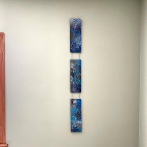 May include: Three rectangular abstract art pieces with blue, teal, and white paint, connected by thin silver wires, hang vertically on a light green wall. The artwork features a textured, layered appearance, creating a modern aesthetic.