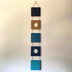 May include: A vertical wall hanging consisting of five square panels connected by silver chains. The panels feature textured surfaces in shades of dark blue, gold, teal, navy blue, and light blue, with two panels containing circular cutouts.