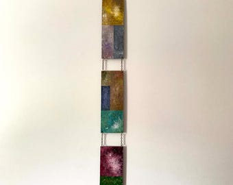 May include: A vertical wall hanging consisting of three rectangular panels connected by metal chains. Each panel features a textured, multi-colored surface with abstract patches of yellow, blue, teal, purple, green, and brown paint.