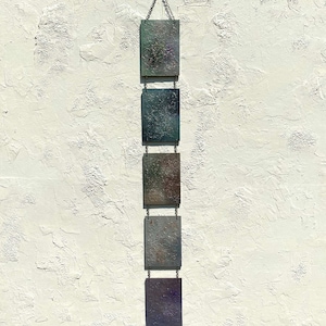 May include: A vertical wall hanging consisting of five rectangular panels connected by metal chains. Each panel features a textured, multi-colored surface with shades of green, blue, brown, and purple, suspended from a single metal ring at the top.