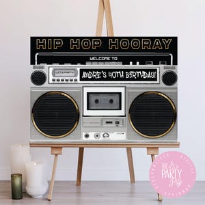 以下が含まれることがあります： 黒い背景に金色の文字で「HIP HOP HOORAY」と書かれた、銀色のブームボックスをテーマにしたパーティーサイン。サインには「WELCOME TO ANDRE'S 40TH BIRTHDAY!」と書かれており、木製のイーゼルに飾られています。レトロなデザイン。