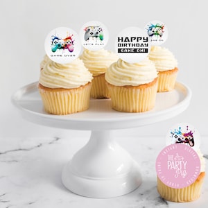 Op de afbeelding: Vier witte cupcakes met witte frosting, bedekt met ronde, kleurrijke, bedrukte papieren cirkels. De cirkels tonen een videogamecontroller met de tekst "Game Over", "Let's Play", "Happy Birthday Game On!" en "Game On".